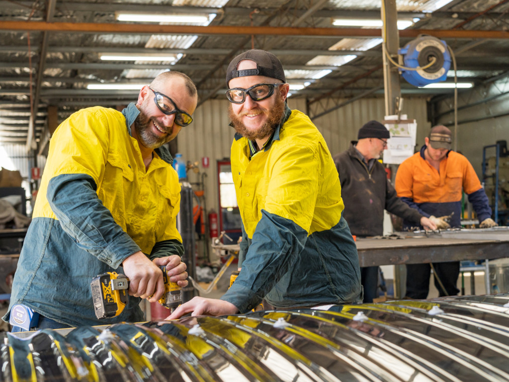 Two sheet metalworkers in yellow workshirts cutting up corrugated metal with an anglegrinder