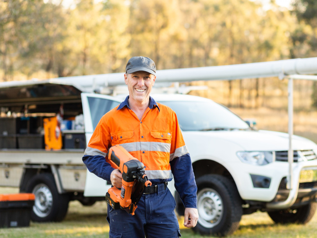 An Aussie tradesman in safety orange work gear carrying a high powered nailgun, standing in front of his work ute.